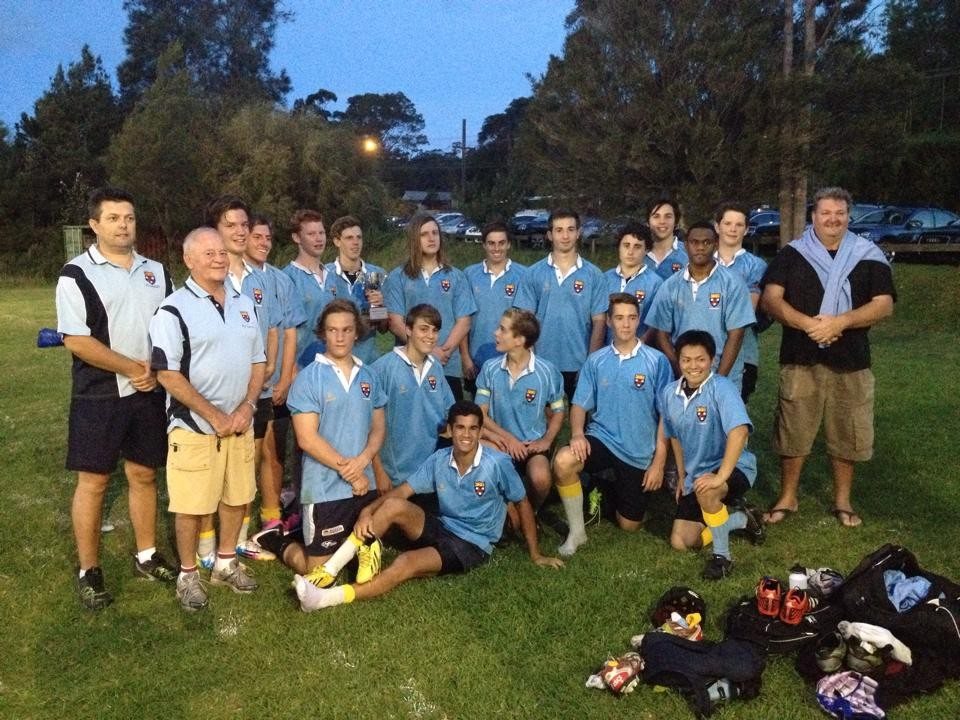A Wakehurst Colts side with a trophy on the field at dusk; Bladesie stands second from left in a club polo and tan shorts.