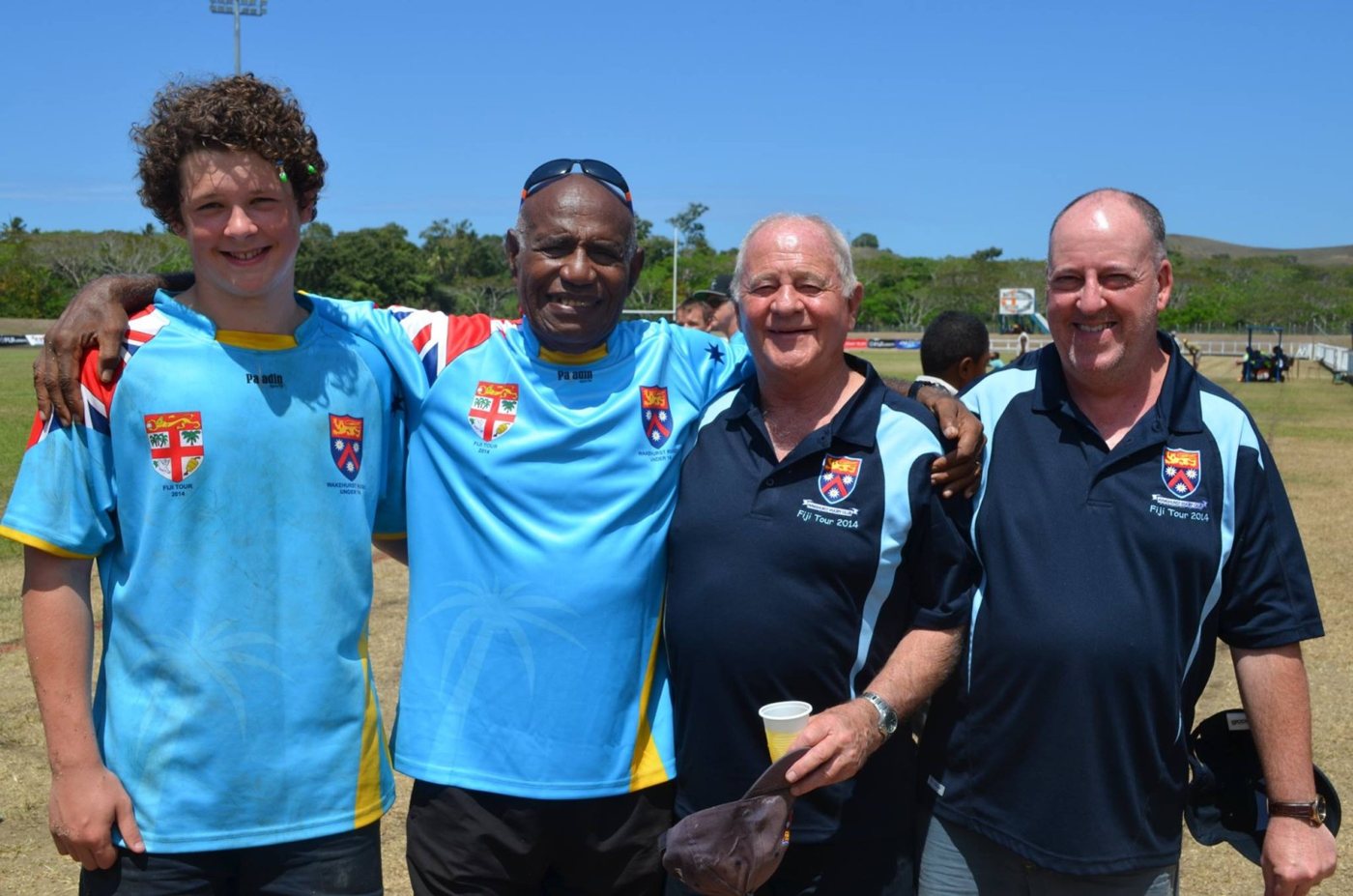 Four Wakehurst tourists at a ground in Fiji in 2014, arms around each other and smiling; Bladesie in a navy Fiji Tour 2014 polo holding a drink.