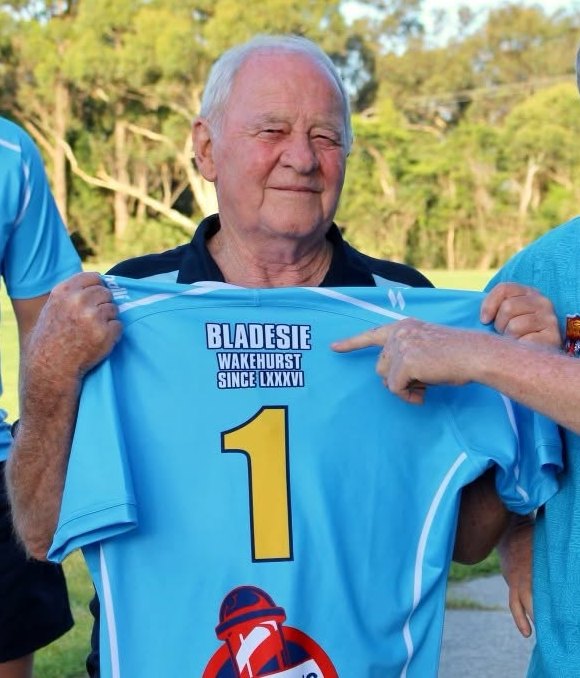 Gareth "Bladesie" Blades smiling as he holds up a sky-blue Wakehurst jersey printed "Bladesie, Wakehurst since LXXXVI" with a big gold number 1.