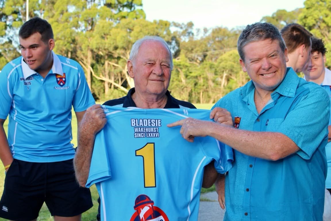 Bladesie at the ground holding up his personalised number 1 Wakehurst jersey, with a club member in a club shirt pointing to his name and a Colts player looking on.