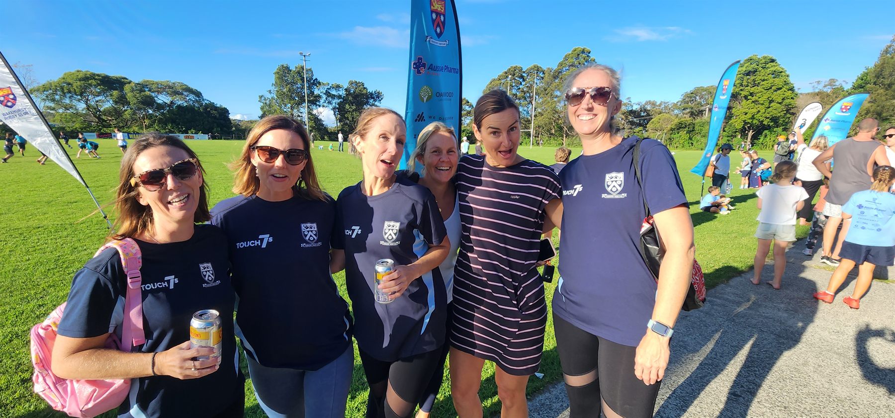 Six Wakehurst women together on the grass beside the club banners on a sunny afternoon at Wakehurst Rugby Park, junior games running behind them.